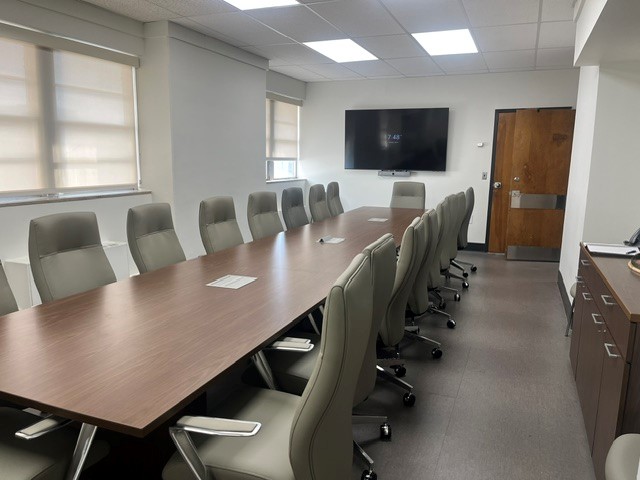 Conference room with long, wooden meeeting table surrounded by padded gray office chairs. In the background, a television monitor is mounted on a wall, and a wooden door is propped open.